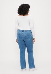 Jean flare bleu en denim, avec poches arrière et taille haute. Associé à un haut blanc à manches longues. Baskets blanches visibles.