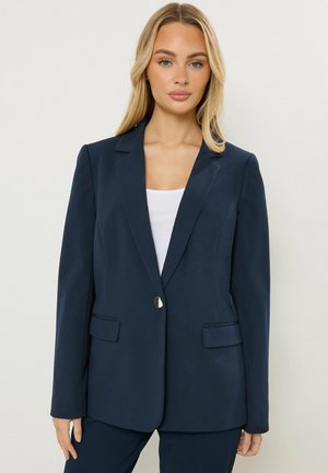 Blonde vrouw draagt een marineblazer over een wit topje, staand tegen een effen lichte achtergrond, met een neutrale uitdrukking naar voren gericht.