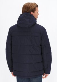 Jachetă puffer de culoare bleumarin, cu glugă, având secțiuni orizontale matlasate și o țesătură texturată. Cusătura din spate este vizibilă, iar designul are o croială lejeră.