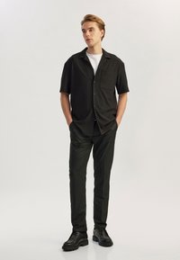 Maglietta nera a maniche corte con taschino, abbinata a pantaloni neri slim-fit e stivali neri con lacci, caratterizzati da una texture liscia e linee pulite.