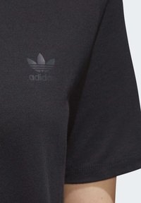Zbliżenie na rękaw czarnej koszulki Adidas z delikatnym, odbijającym światło logo Adidas w kształcie trójlistnej koniczyny na materiale.