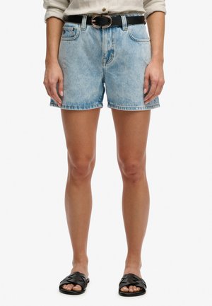 Persoon draagt lichtblauwe denim shorts met voorzakken, een zwarte riem, opgerolde witte blouse en zwarte slidesandalen, staand met ontspannen armen.