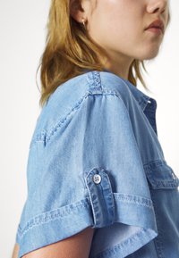 Svetlo modra jeans srajca s kratkimi rokavi, gumbi kot okrasni elementi in vidne šive. Tkanina se zdi gladka in rahlo teksturirana.
