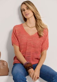 Korallenfarbener Pullover aus Strick mit V-Ausschnitt und kurzen Ärmeln, der ein strukturiertes Muster und goldene Sprenkel aufweist. Akzentuiert mit gestapelten Armbändern.