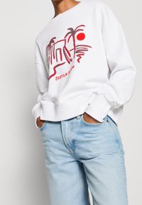 White sweatshirt with red graphic design featuring a sun and palm trees, relaxed fit, soft texture, ribbed cuffs, worn with light blue jeans.