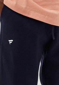 Pantaloni jogger blu Navy in cotone, con vita elasticizzata e dettaglio del logo bianco sulla coscia sinistra, abbinati a una maglietta rosa chiaro.