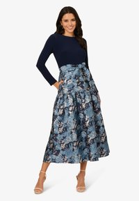 Blumige Maxi-Rock in Blautönen mit Rüschenschichten, kombiniert mit einem marineblauen Langarmoberteil. Der Rock hat einen Bindegürtel und Taschen.