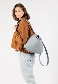 Grauer Leder-Rucksack mit dreieckiger Form, langem verstellbarem Riemen und glatter Textur. Von einer Person auf einer Schulter getragen.