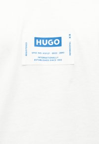 Λευκό ύφασμα με ορθογώνια μπλε και λευκή ετικέτα που αναγράφει "HUGO", μαζί με πληροφορίες για κατοχυρωμένο εμπορικό σήμα και έτος ίδρυσης 1993.