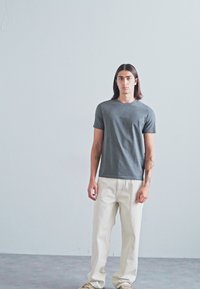 Grå kortermet t-skjorte laget av bomull, kombinert med off-white rette bukser og beige joggesko, stående på en nøytral bakgrunn.