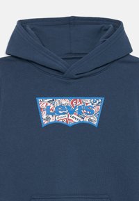 Marineblå hettegenser med en ribbestrikket hette, stort grafisk logo i blått med røde og hvite detaljer, og en jevn tekstur.