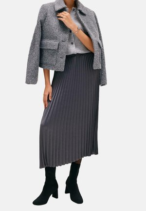 Frau trägt eine graue, strukturierte Jacke über einem grauen Cardigan, einem dunkelgrauen, plissierten Midirock und schwarzen, knöchelhohen Stiefeln mit Absatz.