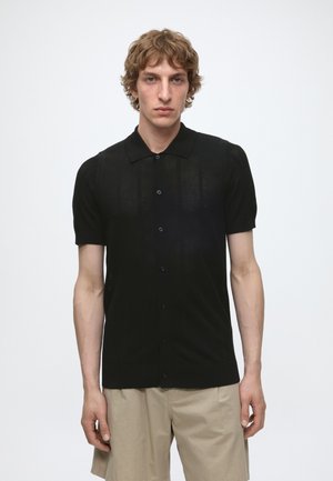 Jonge man met licht golvend haar draagt een zwart, kortmouws, knoopsluitend mesh overhemd en beige korte broek tegen een effen witte achtergrond.