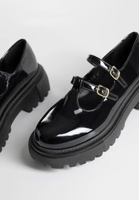 Chaussures Mary Jane en cuir verni noir avec deux boucles réglables, semelle épaisse texturée et design à bout arrondi.