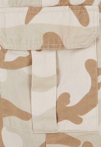Beige camouflage fabric with irregular brown patterns. Features a stitched flap and a visible seam. Smooth texture and durable material.