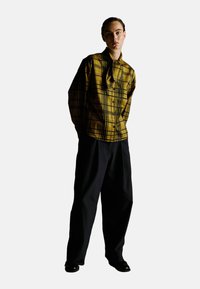 Chemise à carreaux jaunes à manches longues et à design boutonné, assortie à un pantalon large noir et des chaussures sombres. Silhouette simple et moderne.