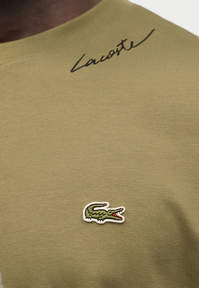 Зехтинено зелена материя с черен курсивен надпис "Lacoste" близо до деколтето и малко зелено бродирано лого на крокодил под него.