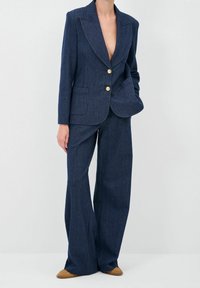 Costume en denim marine, blazer ajusté avec revers prononcés, bouton unique en or, associé à un pantalon à jambes larges, texture lisse, design minimaliste.
