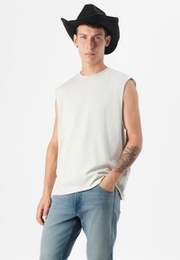 Jonge man in een mouwloos grijs shirt en blauwe jeans, kalm poserend met één hand in zijn zak, getopt met een zwarte cowboyhoed.