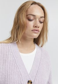 Cardigan lavorato a maglia lilla con scollo a V e motivo testurizzato. Presenta un bottone dorato ed è indossato sopra una camicia bianca.