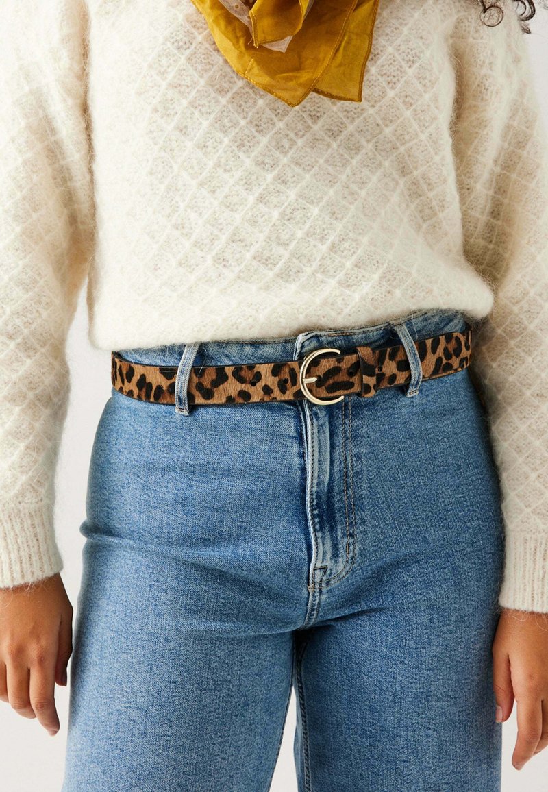 Ceinture à motif léopard avec une boucle circulaire dorée, portée sur un jean bleu taille haute et un pull texturé de couleur crème.
