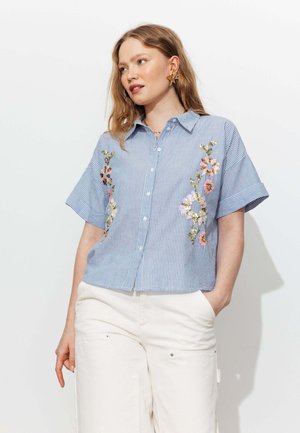 Femme portant une chemise à manches courtes rayée bleu et blanc avec broderies florales, associée à un pantalon blanc, posant devant un fond blanc.