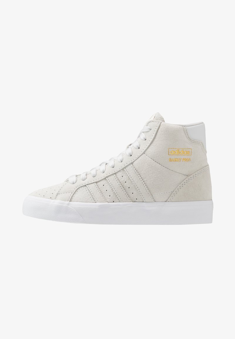 Korkeavartiset beige mokka Adidas -tennarit valkoisella pohjalla, nauhoilla, rei'itetyillä raitapainatuksilla ja kultaisella "adidas BASKET PROFI" -logolla sivussa.
