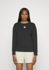 Sweatshirt noir avec un col rond et des manches longues, orné d'un logo brodé circulaire sur la poitrine. Une paire de shorts clairs.