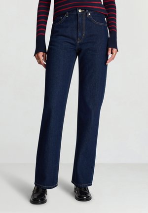 Jeans in denim blu scuro con design a gamba dritta, cuciture visibili e vita alta. Abbinati a scarpe nere e una maglia a righe.