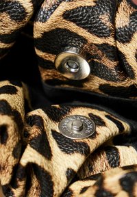 Černý a béžový leopardí vzorovaný kožený materiál s texturovaným povrchem, se stříbrným cvokovým tlačítkem a vyraženým logem na kulaté kovové ozdobě.