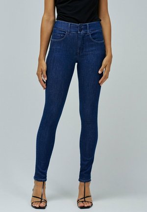 Žena v tmavě modrých skinny jeans s vysokým pasem a dvojitým knoflíkem, doplněná černými sandály na podpatku, před jednobarevným světlým pozadím.