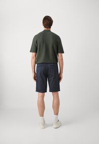 Camicetta a maniche corte verde scuro con una finitura testurizzata, abbinata a pantaloni corti blu navy. Il modello indossa sneakers bianche e calzini alla caviglia.