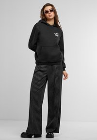 Felpa nera con tasca frontale e grafica bianca, abbinata a pantaloni a gamba larga a righe bianche e nere. Il modello indossa scarpe platform nere.