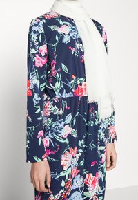Długa sukienka z długim rękawem w kolorze granatowym z żywymi, wielokolorowymi wzorami kwiatowymi. Do tego biała, teksturowana chusta. Elastyczna talia.