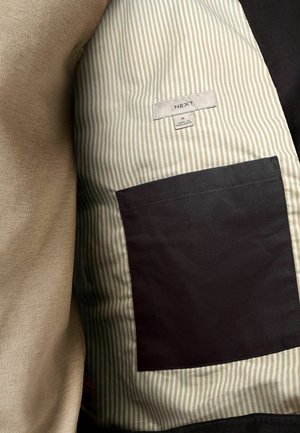 Binnenkant van de jas met beige stof en een gestreept patroon, en een zwarte zak. Label met "NEXT," maat M en details over de stofinhoud.