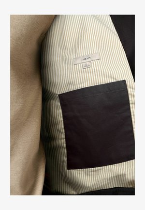 Jakkeinteriør med beige stof med stribet mønster og sort lomme. Etiketten viser "NEXT," størrelse M, og oplysninger om stofindhold.