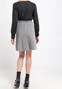 Blouse noire à manches longues avec une texture lisse, assortie à une jupe gris en A. La jupe a un design portefeuille et un aspect tricoté ; bottines noires.