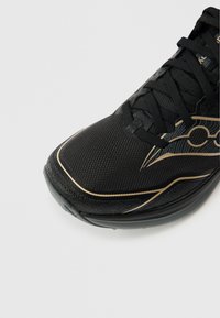 Zwarte hardloopschoen met een structuur van mesh bovenwerk, zwarte veters en een goudkleurig accentontwerp aan de zijkant. Afgeronde zool met een lichte boog.