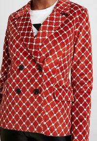 Röd kordflöjels blazer med dubbelknäppning som har ett vitt hjärt- och kedjemönster, svarta knappar och sidofickor.