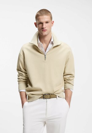 Young man with short hair wearing beige half-zip sweater over white shirt, white pants, and tan belt, hands in pockets, standing against white background.