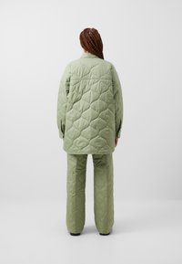 Jachetă verde matlasată și pantaloni asortați dintr-un material texturat cu un model în diamante, având o croială lejeră și detalii de cusătură vizibile.