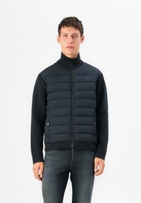 Navy puffer vest with a high collar, stitched sections for insulation, and side pockets, paired with a black long-sleeve shirt and dark jeans.