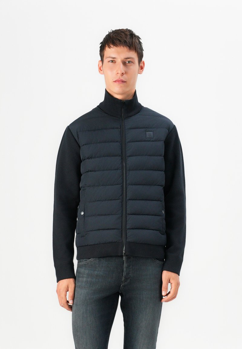 Navy puffer vest with a high collar, stitched sections for insulation, and side pockets, paired with a black long-sleeve shirt and dark jeans.