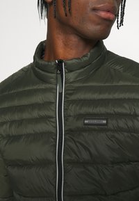 Grön puffjacka med hög krage, framdragkedja och horisontell sömnad. Har ett logopatch på vänster bröst.