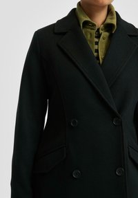 Zwarte dubbelzijdige jas met brede revers, gedragen over een olijfgroen corduroy shirt en een zwart-geel gestreept top.