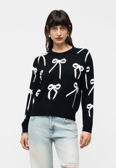 Femme portant un pull noir avec des motifs de nœuds blancs, un jean bleu clair déchiré et des boucles d'oreilles pendantes, debout devant un fond uni.