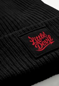 Black knit beanie features a prominent woven label with "Little Devil" in red script, accented by devil horn graphics. Textured fabric.