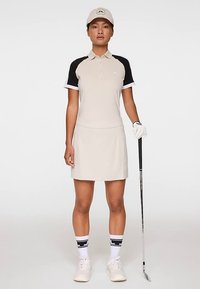 Beige golfpolo med svarta raglanärmar, matchande kjol och vita detaljer. Accessoarer inkluderar keps, handskar, golfklubba och vita sneakers.