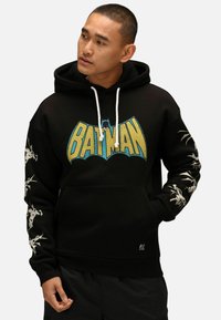 Felpa nera con grafica "BATMAN" in blu e giallo. Presenta un cordino bianco, accenti grafici sulle maniche e tasca a marsupio sul davanti.