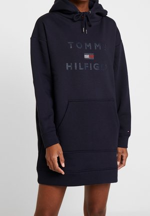 Osoba ubrana w granatową sukienkę z kapturem z napisem "Tommy Hilfiger" i małym logo z przodu, z kieszenią kangurką i sznurkiem przy kapturze.